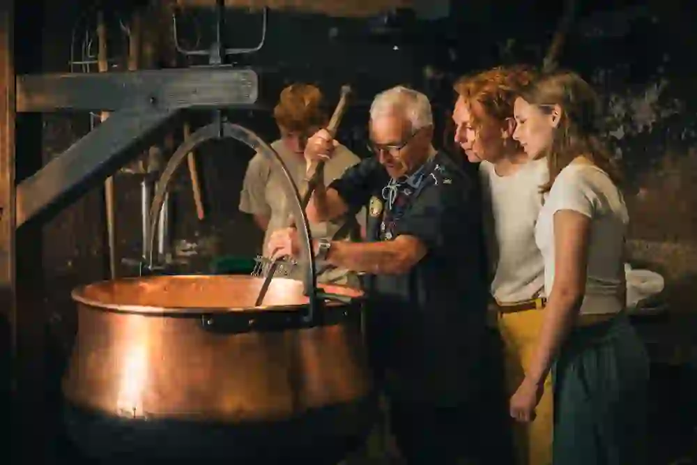 Das Bild zeigt eine traditionelle Käserei, in der ein älterer Mann mit einer Gruppe von Besuchern, darunter zwei junge Frauen und ein junger Mann, an der Käseherstellung arbeitet. Der Mann trägt eine traditionelle Schweizer Tracht und rührt mit einem langen Werkzeug in einem grossen Kupferkessel, der über einer offenen Feuerstelle hängt. Die Besucher beobachten interessiert den Prozess und wirken fasziniert. Die rustikale Umgebung mit dunklen Wänden und hölzernen Balken verleiht dem Bild eine authentische, traditionelle Atmosphäre, typisch für eine Schaukäserei im Emmental.