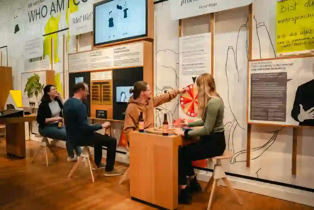 The picture shows an interactive exhibition at the Museum of Communication in Bern. Four people are sitting on stools and taking part in various activities. One person is spinning a wheel on a game table while two others are chatting and holding drinks. On the walls are informative boards and screens covering topics such as culture, language and communication. The atmosphere is relaxed and welcoming, and visitors seem to be actively engaging with the exhibition elements. The exhibits are labelled in German, English and French.