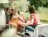 Eine Gruppe von Freunden genießt einen fröhlichen Nachmittag auf einer sonnigen Restaurantterrasse am Fluss. Lachen und gute Laune erfüllen die Luft, während sie ihre Gläser in geselliger Runde erheben. Der idyllische Hintergrund aus grüner Natur und Wasser verstärkt die entspannte und herzliche Atmosphäre.