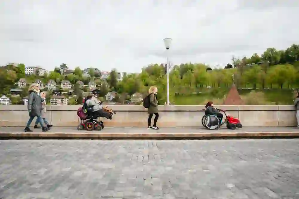 The picture shows people in wheelchairs and companions crossing a bridge, with a picturesque cityscape in the background. The atmosphere is relaxed and everyday, which emphasises the inclusive character of the place.