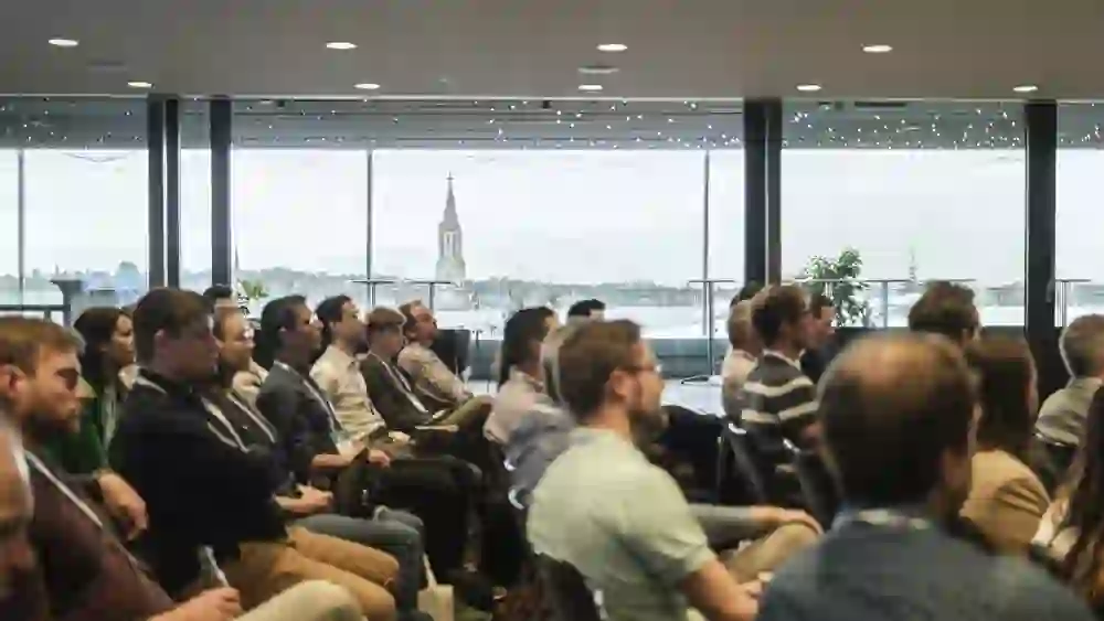 Alt-Text-Vorschlag (mit Stimmung, max. 250 Zeichen):  > Eine konzentrierte Gruppe von Menschen hört in einem modernen Konferenzraum einem Vortrag zu. Durch die Panoramafenster sieht man die Berner Altstadt mit Münster im Hintergrund – eine inspirierende, ruhige Atmosphäre.