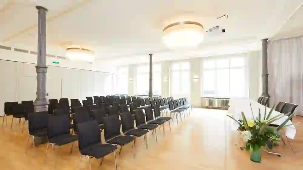 Ein heller, luftiger Saal im Kongresszentrum Kreuz Bern, der mit Reihen von Stühlen für eine Veranstaltung vorbereitet ist. Die große Fensterfront und elegante Beleuchtung schaffen eine Atmosphäre der Erwartung und des professionellen Austausches.