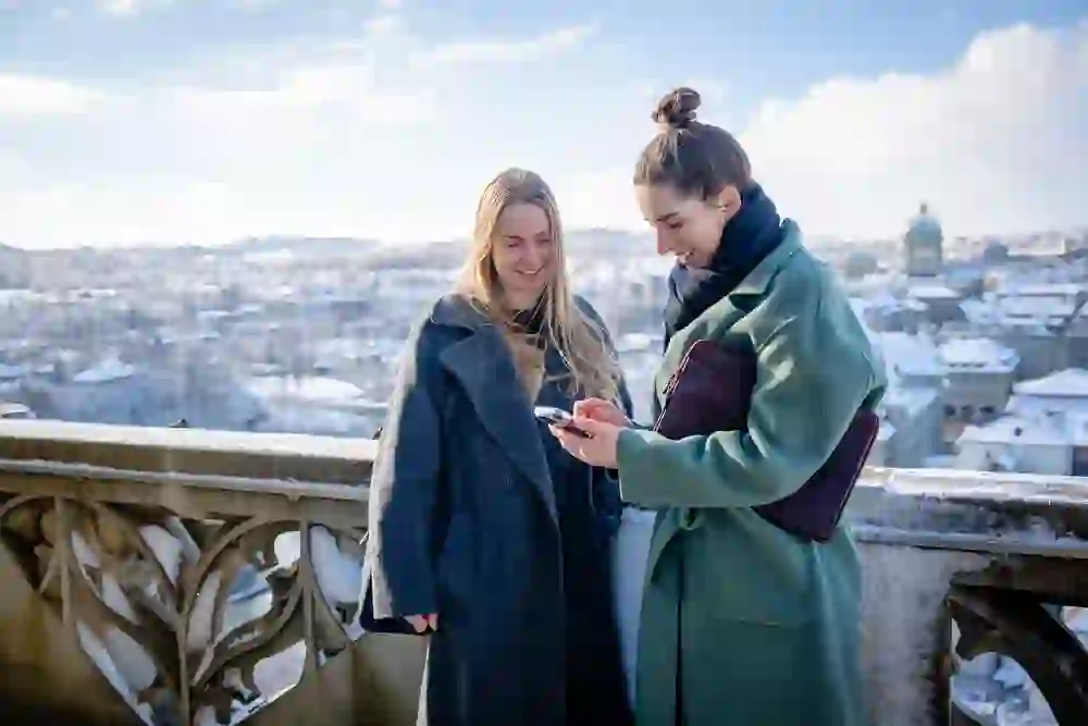 Two women share a serene moment on a terrace overlooking a snowy cityscape, connected by looking at a smartphone.