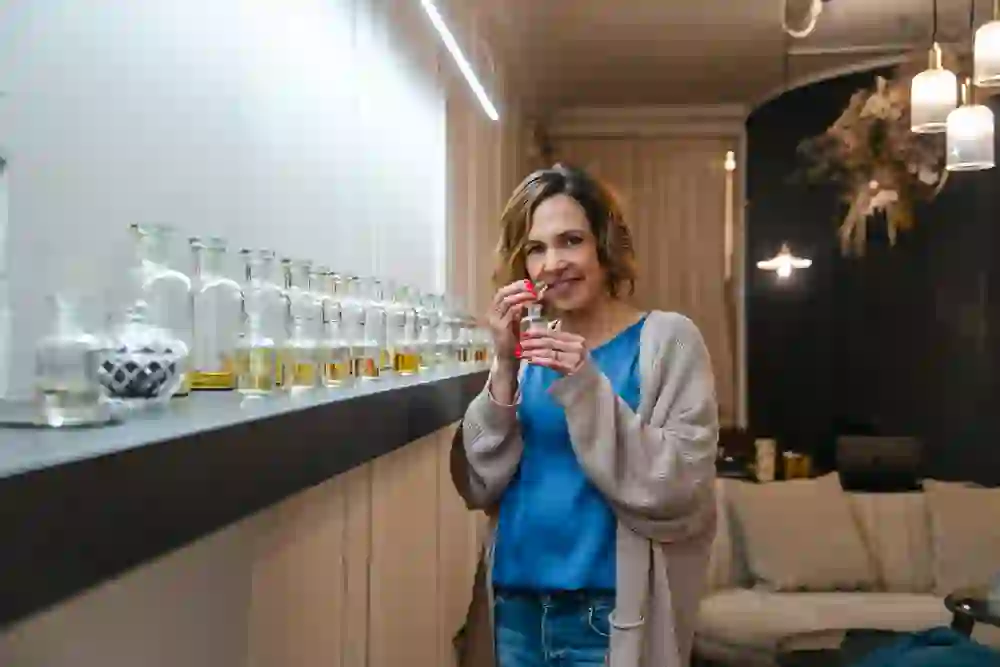 Eine mittelalte Frau riecht an einer Parfumflasche, neben ihr sind diverse Flacons aufgereiht. Sie trägt die blonden Haare zu einem Bob geschnitten. Über dem blauen Shirt trägt sie eine beige Strickjacke, dazu blaue Jeans.