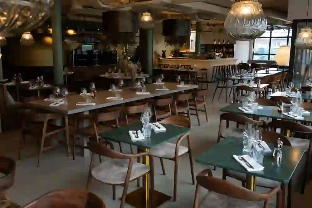 L'image montre l'intérieur d'un restaurant aménagé avec style. La salle est équipée de plusieurs tables et chaises en bois préparées pour les clients. Les tables sont recouvertes d'assiettes, de verres et de couverts pour le repas. Il y a aussi bien de longues tables pour les groupes que des tables plus petites pour deux personnes. De grandes lampes décoratives sont suspendues au-dessus des tables, ce qui crée une atmosphère chaleureuse. En arrière-plan, on aperçoit le bar du restaurant, équipé de différentes boissons et d'une cuisine moderne.