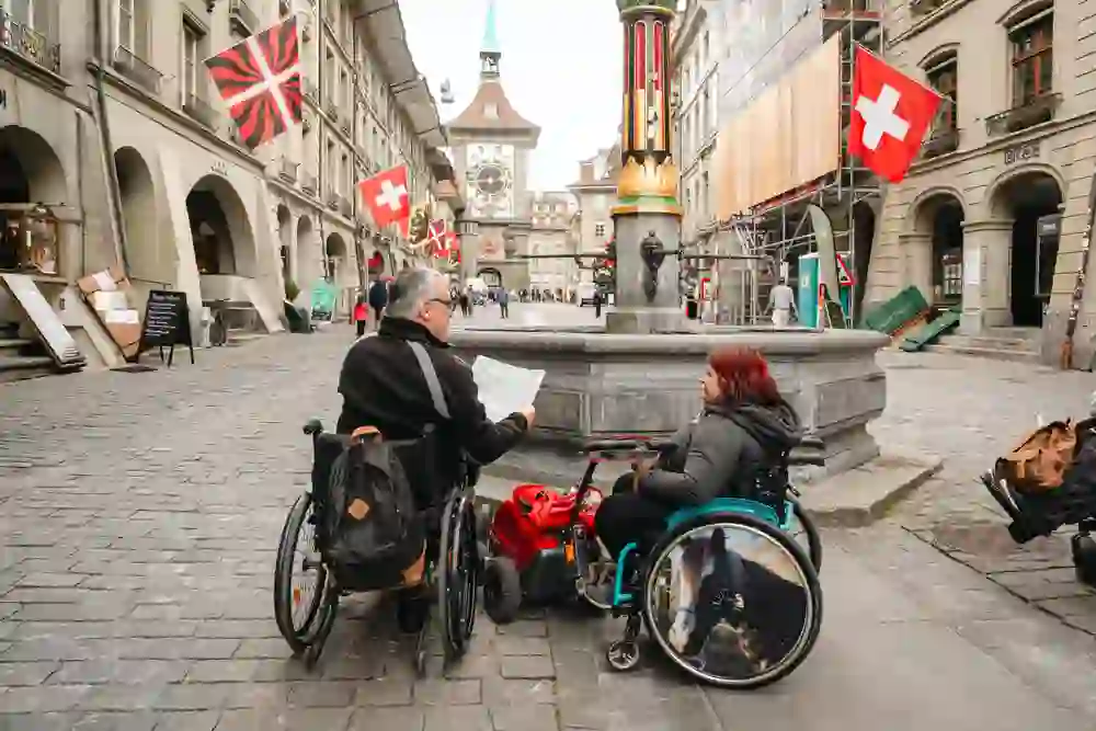 L'image montre des personnes en fauteuil roulant explorant le centre-ville animé, avec son architecture historique et ses drapeaux suisses. L'atmosphère est vivante et inclusive, avec un sentiment de communauté et d'accessibilité.