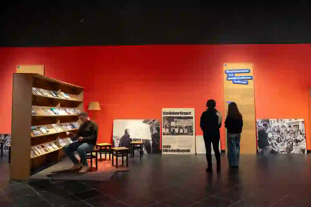 Das Bild zeigt eine moderne Ausstellung in einem Museum mit dem Thema Menschenrechte und gesellschaftlicher Aufbruch. Eine rote Wand dient als Hintergrund für mehrere Informationstafeln, historische Fotografien und Plakate, die auf Protestbewegungen und soziale Kämpfe hinweisen, darunter ein Plakat mit der Aufschrift „Solidarität mit Hindelbank“ und ein weiteres, das sich auf das Stimmrecht für Frauen bezieht.  Links im Bild sitzt eine Person an einem Lesebereich mit einem Bücherregal, das mit verschiedenen Publikationen bestückt ist. Zwei weitere Personen betrachten interessiert die Ausstellungstafeln. Die Szene vermittelt eine nachdenkliche und reflektierte Atmosphäre, in der sich Besucher:innen mit historischen und gesellschaftlichen Themen auseinandersetzen.