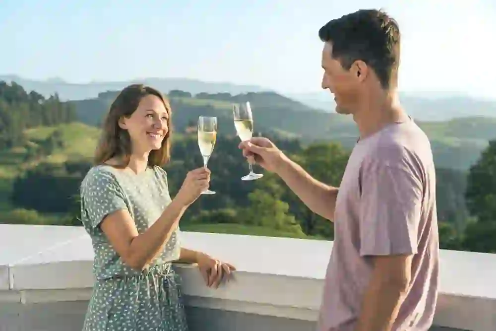 Sur la photo, une femme et un homme portent un toast avec des flûtes à champagne. Ils se tiennent dehors devant un paysage pittoresque de douces collines et de forêts. Le soleil brille et tous deux se sourient, ce qui crée une ambiance chaleureuse et joyeuse.