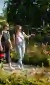 Two women stroll through a blooming rose garden, one smiling and pointing to something in the distance. The atmosphere is peaceful and relaxed, surrounded by colourful flowers and bright sunshine. It seems like a carefree moment in nature.