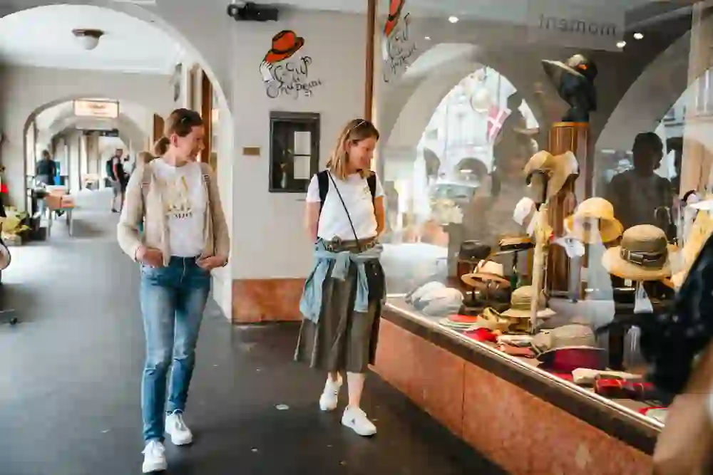 Das Bild zeigt zwei Frauen, die entspannt an einem Schaufenster mit Hüten in der Berner Altstadt vorbeischlendern. Sie wirken fröhlich und gelassen, während sie die Auslagen betrachten. Die Atmosphäre ist locker und gemütlich, voller urbanem Charme und Freude am Entdecken.
