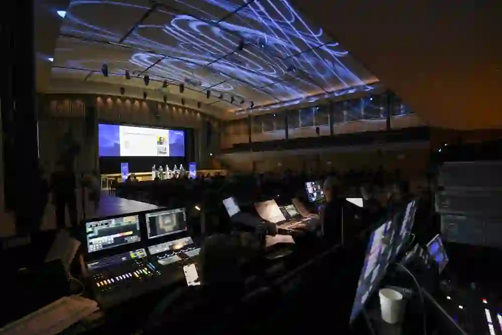 L'immagine mostra un centro di controllo tecnologico per eventi professionali durante un congresso o una conferenza. In primo piano si vede l'area tecnica con diversi schermi, console per l'illuminazione e per il suono, dove si concentrano gli addetti. Sullo sfondo si vede un palco con altoparlanti e un grande schermo di proiezione su cui vengono visualizzate le presentazioni. Il design dell'illuminazione proietta eleganti motivi blu sul soffitto, conferendo alla sala un'atmosfera moderna e di alta qualità. L'atmosfera è concentrata e professionale.