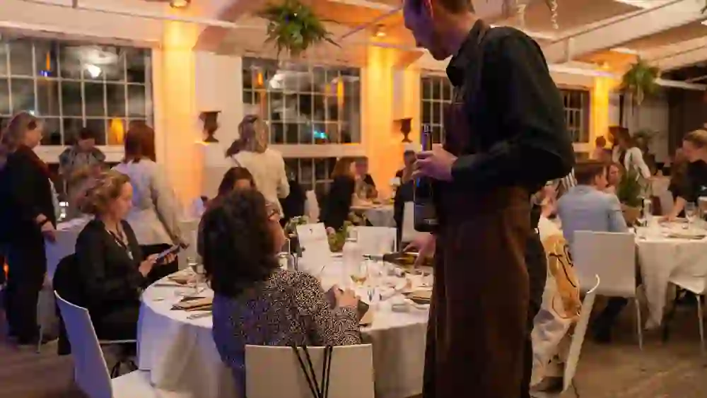 In einem stilvoll dekorierten Saal mit warmem Licht genießen die Gäste ein festliches Dinner. Ein Kellner serviert Wein am Tisch „BERNEXPO“, während die Gäste plaudern und sich sichtlich wohlfühlen – die Stimmung ist entspannt und festlich.