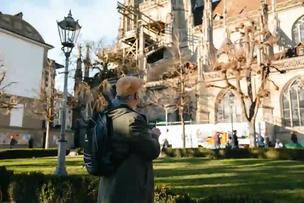 Un uomo anziano con le cuffie segue un tour audio di Berna. Si ferma davanti all'imponente cattedrale, ascolta con attenzione l'audioguida e scopre la città in modo personale e decelerato: curioso, attento e completamente immerso nel momento.