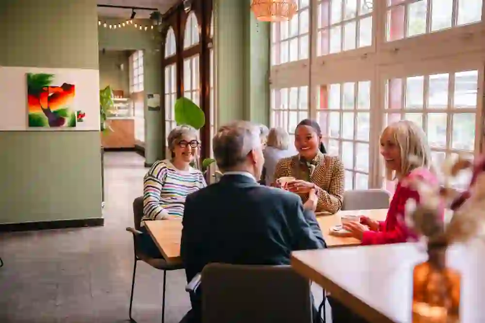 Dans l'orangerie baignée de lumière, quatre personnes sont assises en toute décontraction autour d'un thé et d'une conversation animée. Entourées de plantes, d'art et d'une atmosphère chaleureuse, elles vivent un moment de complicité, de sérénité et de véritable joie de vivre.        4o