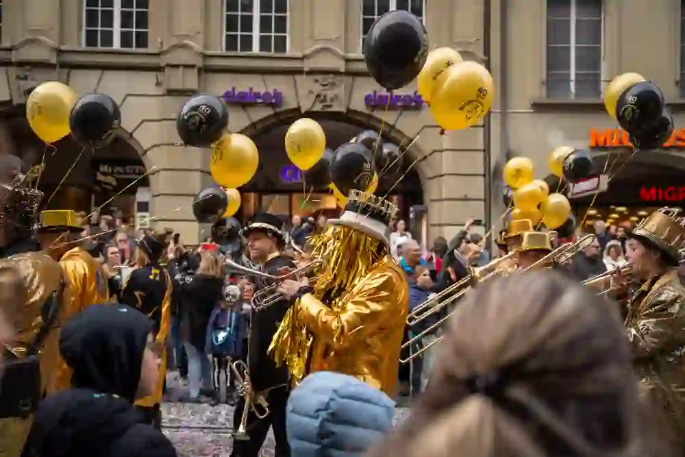 L'immagine mostra un gruppo di Guggenmusik al Carnevale di Berna, che suona musica in vistosi costumi dorati. Alcuni dei musicisti indossano cappelli e maschere scintillanti, che sottolineano l'atmosfera di festa. Al gruppo sono legati dei palloncini neri e oro con la scritta "40 anni", a indicare un anniversario. Sullo sfondo, numerosi spettatori assistono allo spettacolo colorato, mentre i coriandoli decorano la strada e creano un'atmosfera vivace.