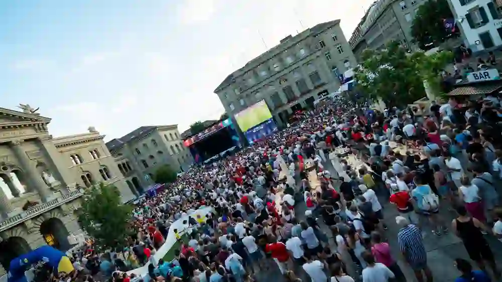 L'image montre une foule immense sur la Place fédérale - la foule suit avec impatience le match de l'équipe nationale féminine suisse.  En arrière-plan, on aperçoit la grande roue et le Palais fédéral.