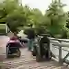 L'image montre des personnes en fauteuil roulant et des accompagnateurs sur un pont au-dessus d'une rivière, entourés d'une verdure luxuriante. L'ambiance est détendue et amicale, tandis qu'ils profitent de la nature et interagissent les uns avec les autres.