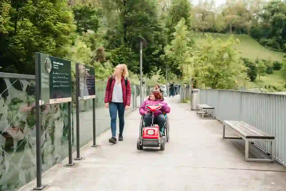 Das Bild zeigt eine Frau, die eine andere Frau im Rollstuhl durch einen Park schiebt, vorbei an Informationsdisplays. Die Atmosphäre ist entspannt und einladend, mit einem Hintergrund aus üppigem Grün und einem friedlichen Weg.