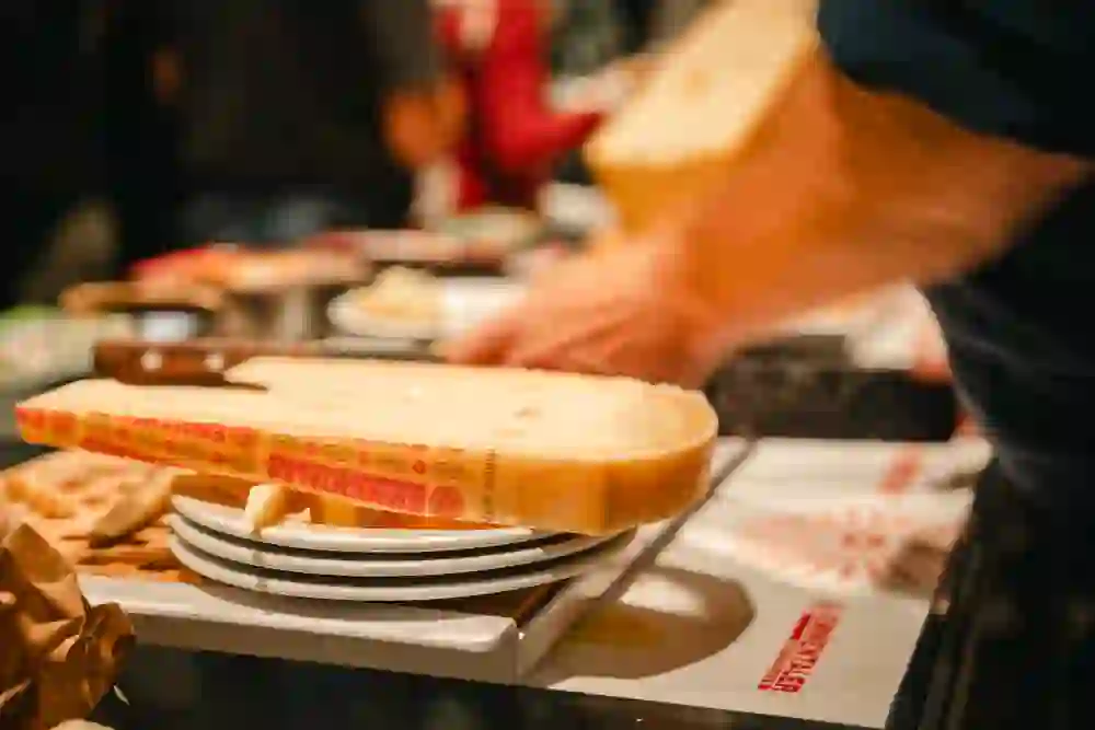 Das Bild zeigt eine Szene mit halbiertem Emmentaler Käse, der auf mehreren Tellern ruht – vermutlich bei einer Käseverkostung oder einem Raclette-/Fondue-Event. Es ist deutlich das rote „Emmentaler Switzerland“-Label auf der Käserinde zu erkennen, was auf Schweizer Herkunft und einen geschützten Ursprungsstatus (g.U.) hinweist.