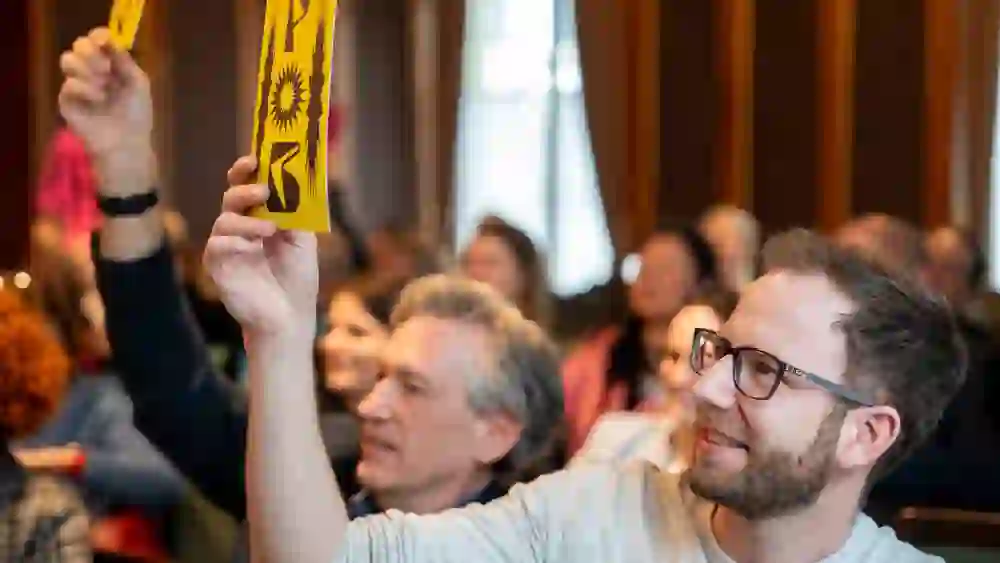 Zwei Männer heben gelbe Karten bei einer Versammlung; der Vordere schaut erwartungsvoll, die Atmosphäre ist engagiert und positiv.