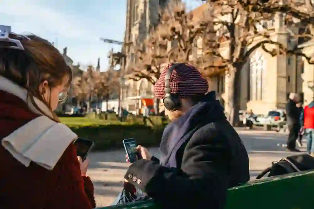 Zwei Personen sitzen entspannt auf einer Bank in der Wintersonne, vertieft in ihre Audiotour. Zwischen historischen Mauern und kahlen Bäumen wird Geschichte lebendig – eine stille Entdeckungsreise mit offenem Blick und offenen Ohren.