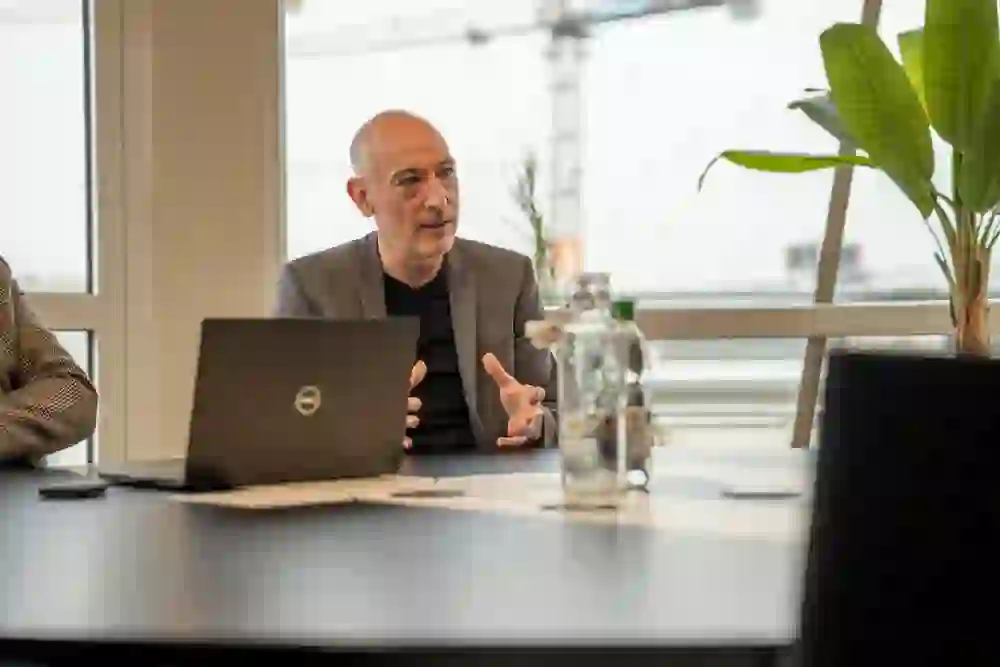 Un homme à l'air sérieux gesticule pendant un entretien, concentré et engagé, dans un bureau baigné de lumière avec un décor urbain en arrière-plan.