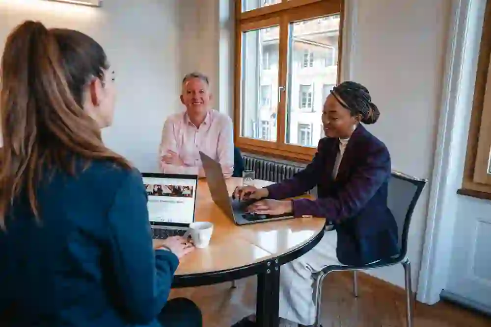 Trois personnes en train d'échanger des idées dans une salle de réunion moderne à Berne. Une lumière du jour claire, une atmosphère ouverte et une collaboration concentrée transmettent professionnalisme, inspiration et une ambiance d'affaires motivante.