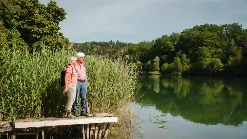 Ein älteres Paar steht eng umschlungen auf einem kleinen Holzsteg am ruhigen See, umgeben von sattem Grün. Der Blick schweift verträumt über das spiegelglatte Wasser. Die Szene strahlt Geborgenheit, Harmonie und die Freude an gemeinsamen Momenten in der Natur aus.