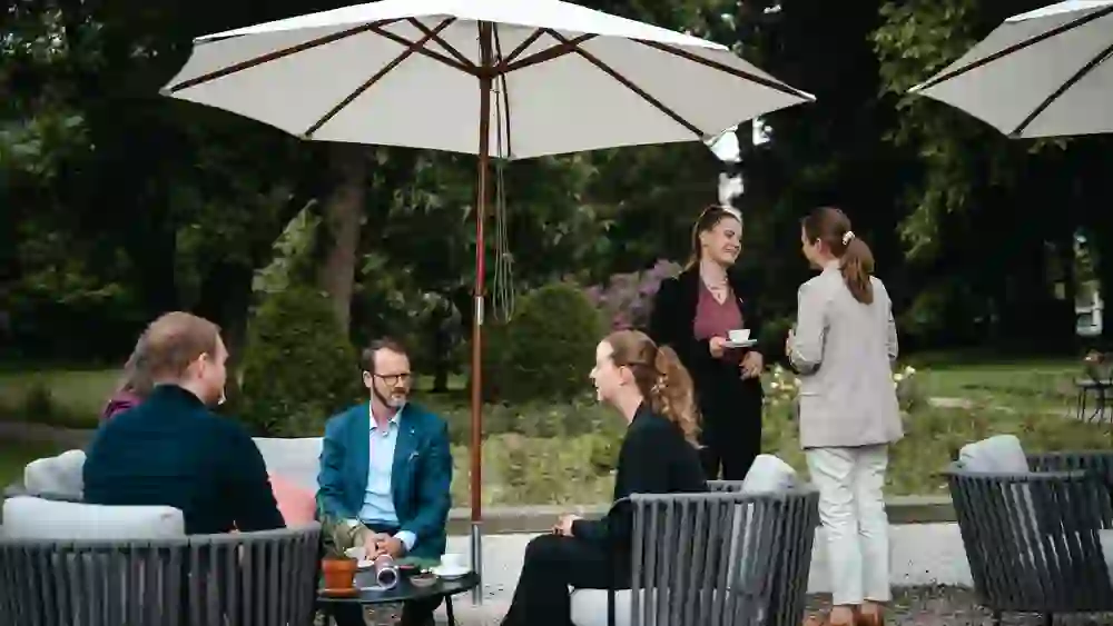 Das Bild zeigt eine Gruppe von Personen, die auf einer Terrasse unter Sonnenschirmen sitzen und sich unterhalten. Einige trinken Kaffee, die Atmosphäre ist entspannt und freundlich, mit einer professionellen, aber lockeren Stimmung im Grünen.
