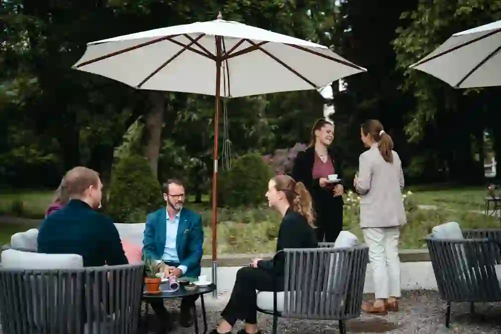 The picture shows a group of people sitting on a terrace under parasols and chatting. Some are drinking coffee, the atmosphere is relaxed and friendly, with a professional but informal mood in a green setting.