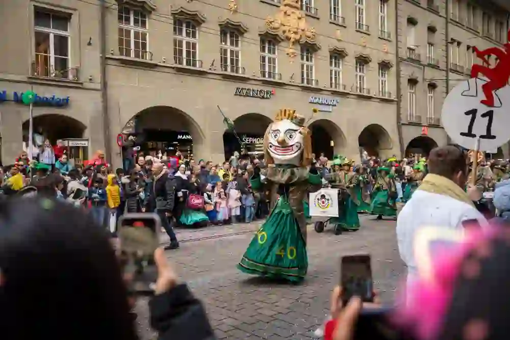 L'immagine mostra una scena del Carnevale di Berna con una figura in un vistoso costume verde e una maschera sovradimensionata dal design umoristico. Sullo sfondo, numerosi spettatori, tra cui famiglie con bambini, si trovano lungo la strada e osservano il corteo. L'atmosfera festosa è sottolineata da dettagli colorati e coriandoli.