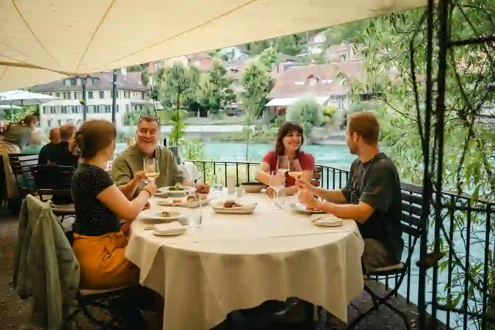 Eine fröhliche Gruppe von Freunden sitzt entspannt auf der Terrasse eines Restaurants direkt am Fluss. Sie genießen gemeinsam ein Essen, lachen und stoßen mit Weingläsern an. Die idyllische Flusslandschaft und die gemütliche Atmosphäre verstärken das Gefühl von Freude und Zusammengehörigkeit.