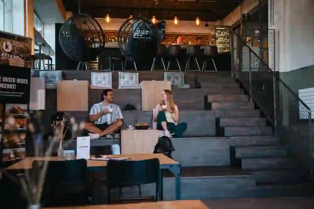 The picture shows the Kraftakt bistro in Stettlen. Two people are sitting relaxed on the wooden steps, drinking coffee and chatting cordially. The warm lighting and modern design create an inviting, cosy atmosphere.