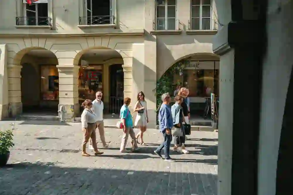 Un gruppo colorato passeggia di buon umore nel centro storico di Berna. L'atmosfera estiva è rilassata, la luce è calda. Conversazioni e sorrisi caratterizzano la scena davanti ai portici storici e alle invitanti vetrine dei negozi.