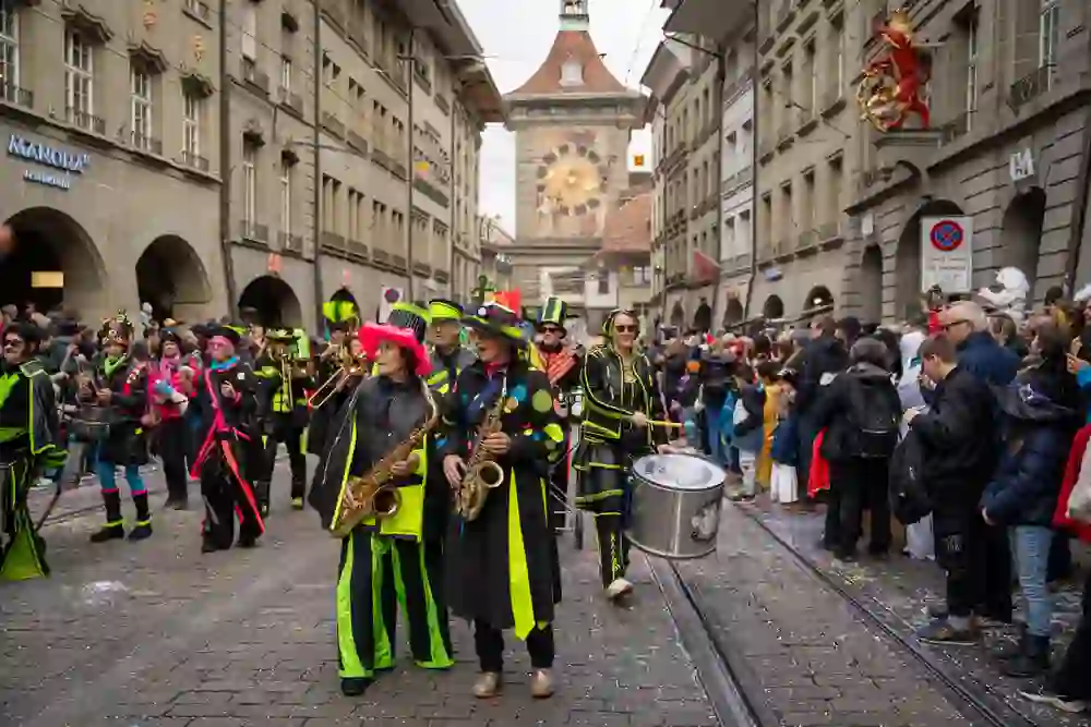 L'immagine mostra un colorato gruppo di Guggenmusik durante il Carnevale di Berna. I musicisti indossano costumi dai colori sgargianti con accattivanti colori al neon mentre sfilano per le strade del centro storico con sassofoni, tamburi e altri strumenti. Sullo sfondo si vede la torre Zytglogge, simbolo di Berna, e una folla di spettatori entusiasti che assistono allo spettacolo. I coriandoli a terra e l'atmosfera festosa sottolineano l'atmosfera vivace.
