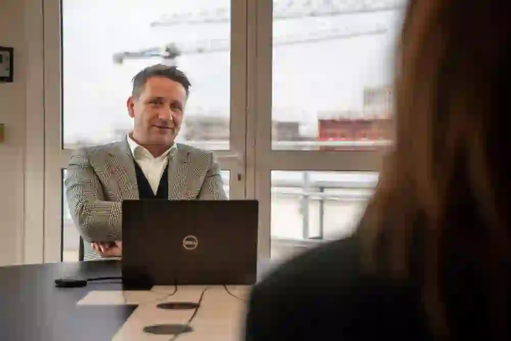 Un homme vêtu d'une élégante veste à carreaux est assis à table, souriant, avec en face de lui un interlocuteur invisible. Attitude ouverte et attentionnée, ambiance communicative et positive.