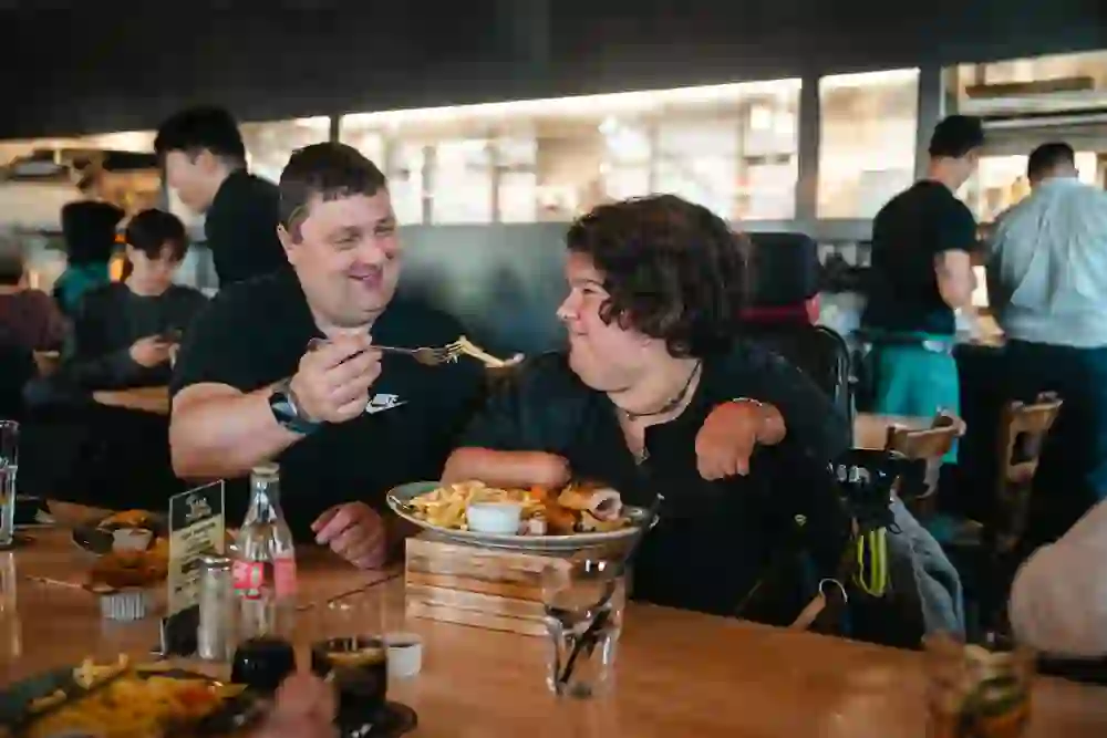 A man lovingly feeds a woman in a wheelchair in a cosy restaurant. They both laugh heartily, their gazes full of warmth. The atmosphere is warm and familiar, the shared meal a moment full of joy and connection.