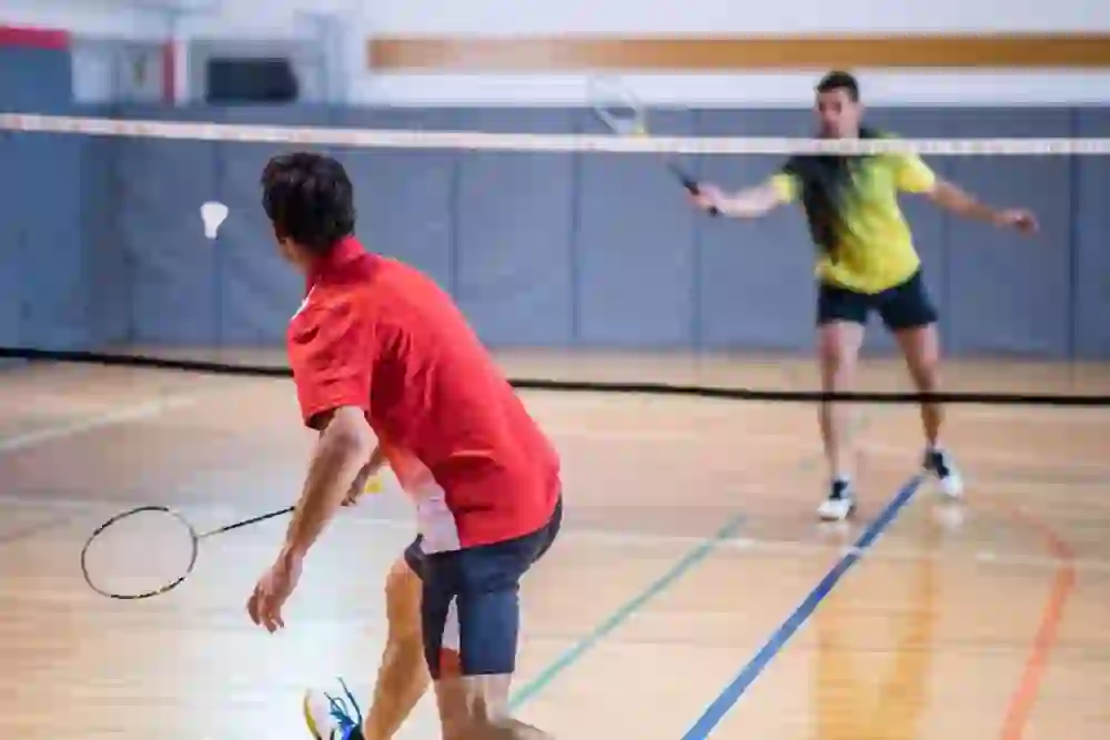 The picture shows a badminton game in a hall. Two players are in the game, one at the net about to hit the shuttlecock while the other prepares for the next move.