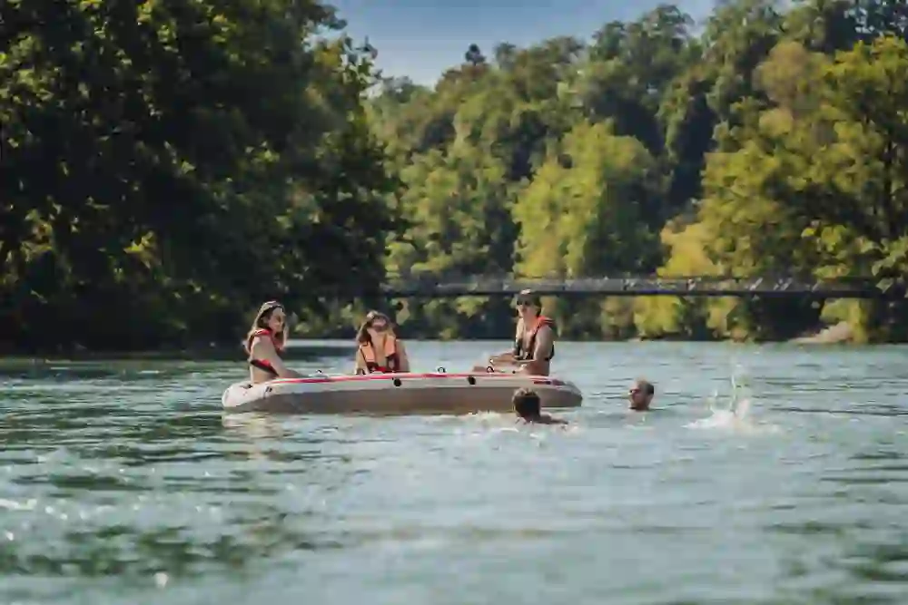 Eine Gruppe lacht und genießt eine Bootsfahrt auf einem Fluss, umgeben von grüner Natur. Eine Person schwimmt nebenbei, was auf einen entspannten und vergnüglichen Sommertag hindeutet.