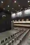 Das Bild zeigt ein modernes Theater mit schwarzen Vorhängen und einer geräumigen Bühne, umgeben von Reihen grauer Sitze. Die minimalistische und elegante Gestaltung schafft eine fokussierte und professionelle Atmosphäre für Aufführungen.