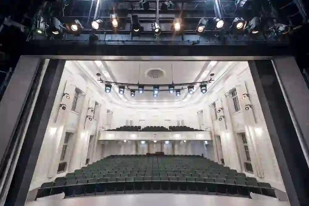 The picture shows an empty theatre stage from the perspective of an actor, surrounded by magnificent green seats and elegant white ornaments. The lighting sets the stage spectacularly, creating anticipation for the curtain call.