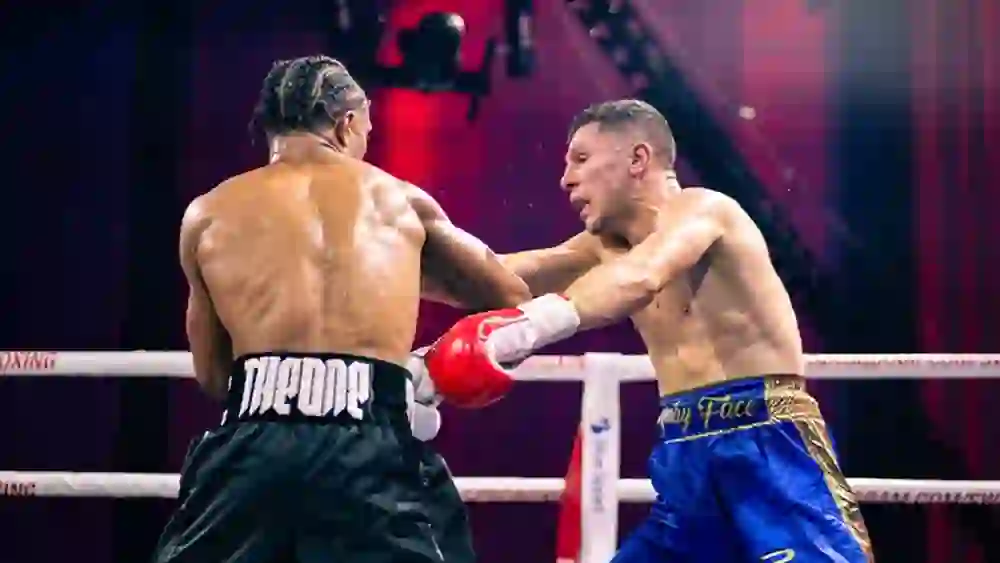 L'immagine mostra un intenso incontro di boxe sul ring. Due pugili danno il meglio di sé, il sudore scorre a fiumi, la tensione è palpabile. L'illuminazione rossa intensifica l'atmosfera drammatica. La determinazione e l'adrenalina la fanno da padrone, il pubblico si emoziona a ogni pugno.