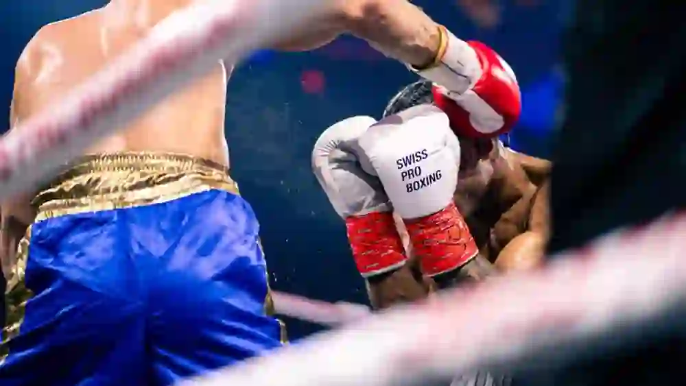 L'immagine mostra un incontro di boxe nella fase decisiva. Un pugile si nasconde dietro i guantoni mentre l'avversario colpisce con forza. Il sudore e la tensione sono nell'aria, il pubblico può percepire l'intensità di ogni movimento. Concentrazione e volontà allo stato puro.