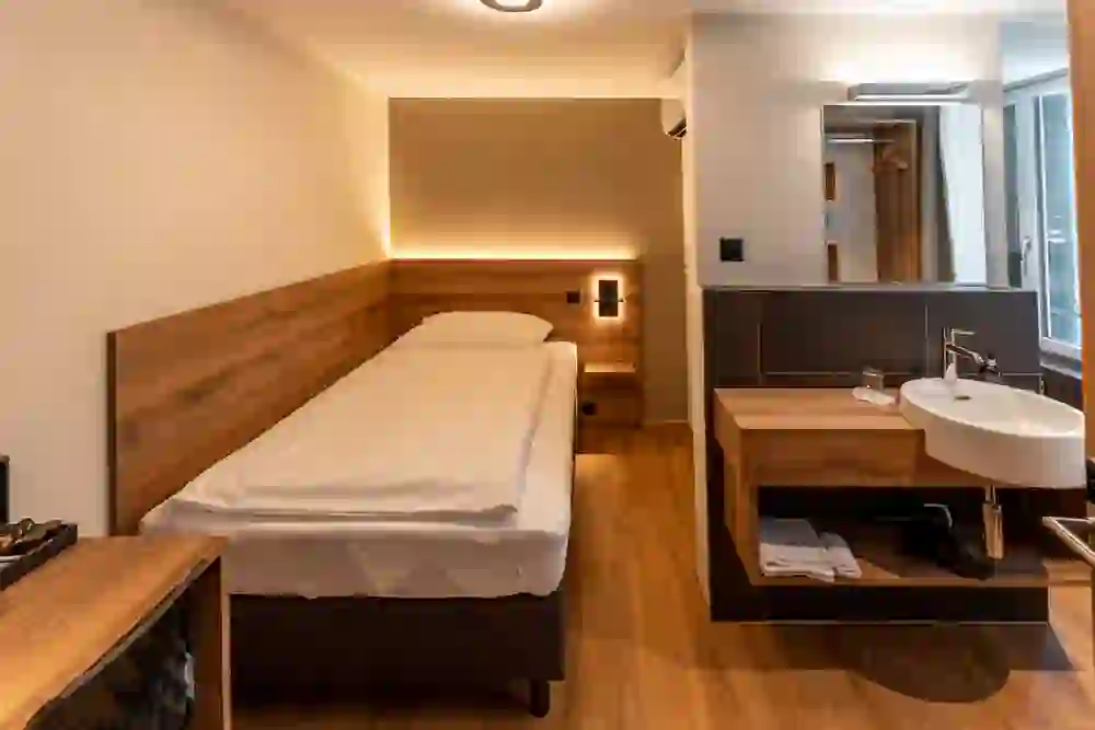 Das Bild zeigt ein kleines, aber gemütlich wirkendes Einzelzimmer. Ein Einzelbett mit einem hellen Laken ist an der Wand platziert, darüber eine indirekte Beleuchtung. Rechts befindet sich ein Waschbecken mit einem großen Spiegel darüber, das scheinbar in den Raum integriert ist, und dahinter ein Fenster. Die Kombination aus warmen Holztönen und sanfter Beleuchtung schafft eine einladende und ruhige Atmosphäre. Es scheint ein modernes und praktisches Design für eine Unterkunft zu sein.