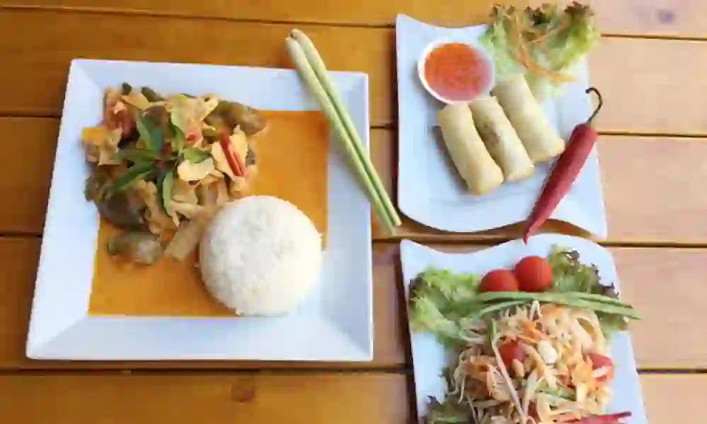 Auf dem Bild ist ein köstliches thailändisches Gericht zu sehen: ein Teller mit duftendem Reis und würzigem Curry, begleitet von knackigen Frühlingsrollen mit Dip-Sauce und einem frischen Papayasalat. Die lebhaften Farben und die Vielfalt der Aromen erzeugen ein Gefühl von exotischem Genuss und Vorfreude auf ein authentisches Geschmackserlebnis.