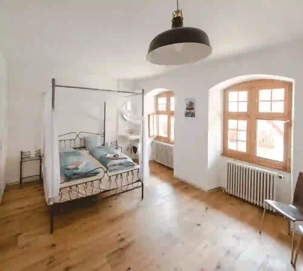 Das Bild zeigt ein helles, einladendes Schlafzimmer mit natürlichem Licht im Schloss Laupen, das durch grosse Holzfenster strömt. Das Zimmer vermittelt eine friedliche, behagliche Atmosphäre mit einem eleganten Himmelbett, weichen blauen Akzenten und einem warmen Holzboden. Das Schloss Laupen liegt an der E-Bike Route Zeitschlaufe Laupen.