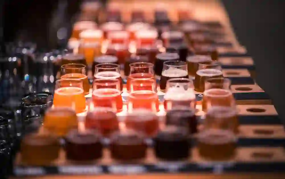 La foto presenta un'impressionante collezione di campioni di birra, ordinatamente disposti e pronti per la degustazione. La tavolozza dei colori spazia dal giallo pallido all'ambra e ai rossi intensi, sottolineando la varietà delle birre offerte. La scena è ambientata in una stanza poco illuminata e l'attenzione si concentra sui bicchieri pieni, creando l'aspettativa di un'esperienza ricca di sapori.