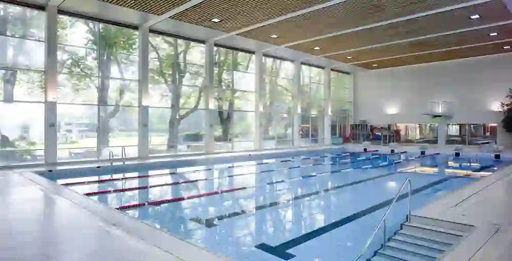 Das Bild zeigt ein modernes Hallenbad mit großen Fenstern, durch die helles Tageslicht strömt. Die klaren Linien und die spiegelnde Wasseroberfläche schaffen eine ruhige, einladende Atmosphäre. Die leeren Bahnen und der stille Innenraum vermitteln ein Gefühl von Sauberkeit und Entspannung.