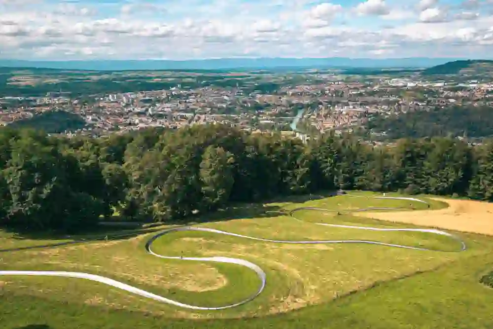 Un enfant descend la pente sur le téléphérique circulaire, entouré de prairies et d'arbres verdoyants. En arrière-plan s'étend l'impressionnant paysage urbain de Berne avec des montagnes à l'horizon.