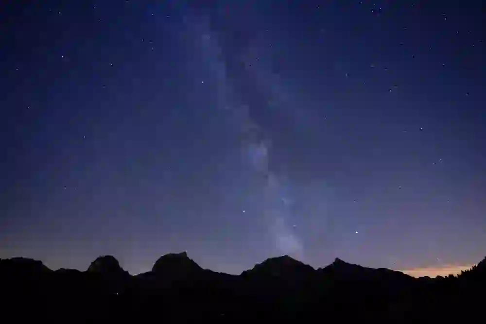 The photo captures a breathtaking night sky in the Gantrisch Nature Park, dominated by the sparkling Milky Way stretching across the panorama of a mountain range. The starry sky is clear and vast, full of countless stars that give an overwhelming impression of the vastness of the universe. The dark contours of the mountains provide a stark contrast to the vivid celestial phenomenon and ground the celestial spectacle.